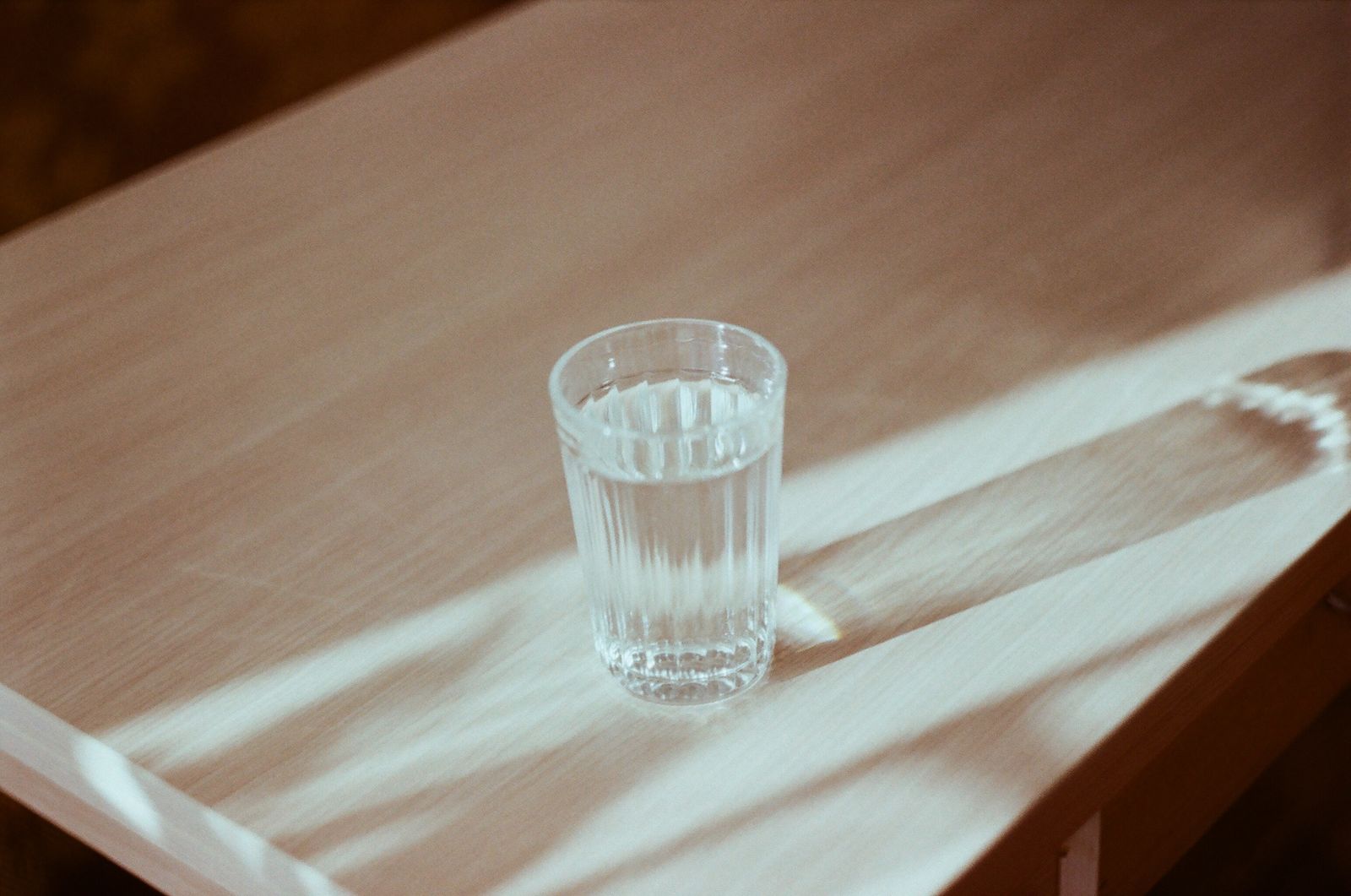 Wasserglas auf einem hellen Holztisch, Fensterlicht wirft weiche Lichtstreifen und Schatten