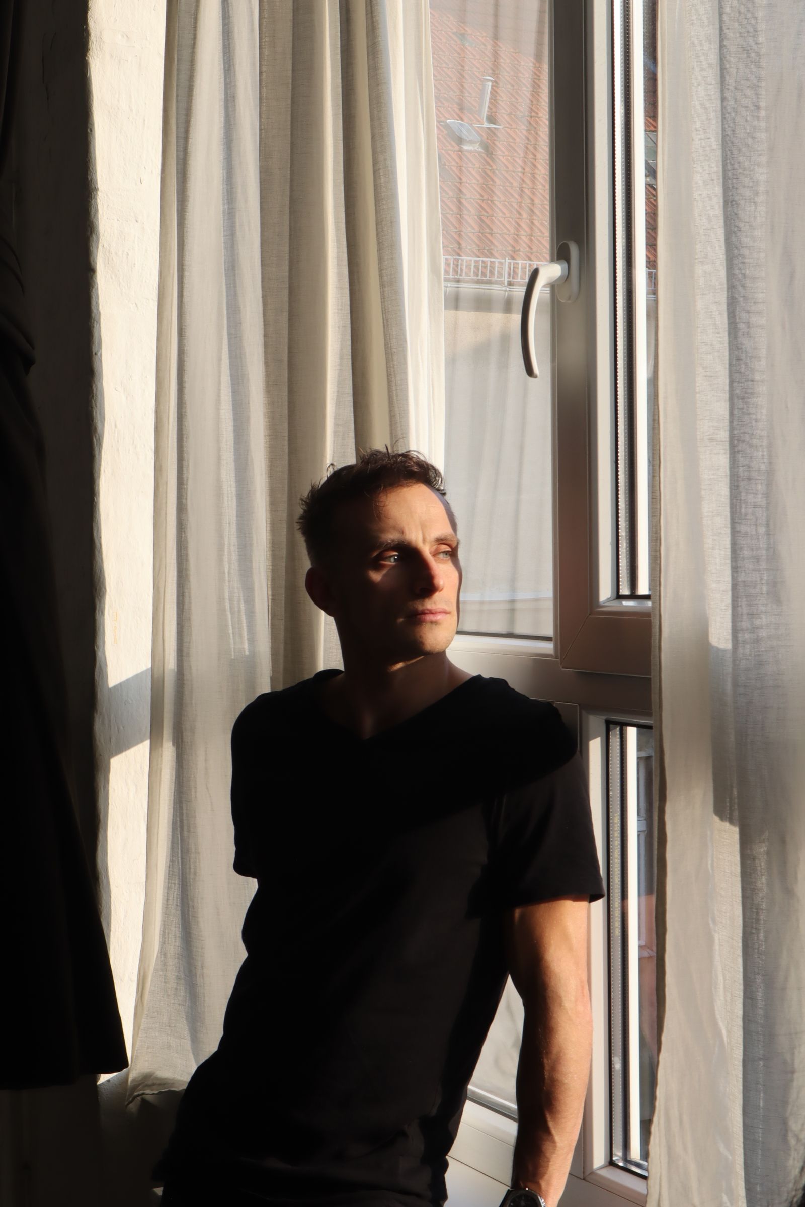 Mann im schwarzen T-Shirt sitzt am Fenster einer Altbauwohnung, warmes Abendlicht fällt durch helle Vorhänge, sein Blick geht nachdenklich nach draußen