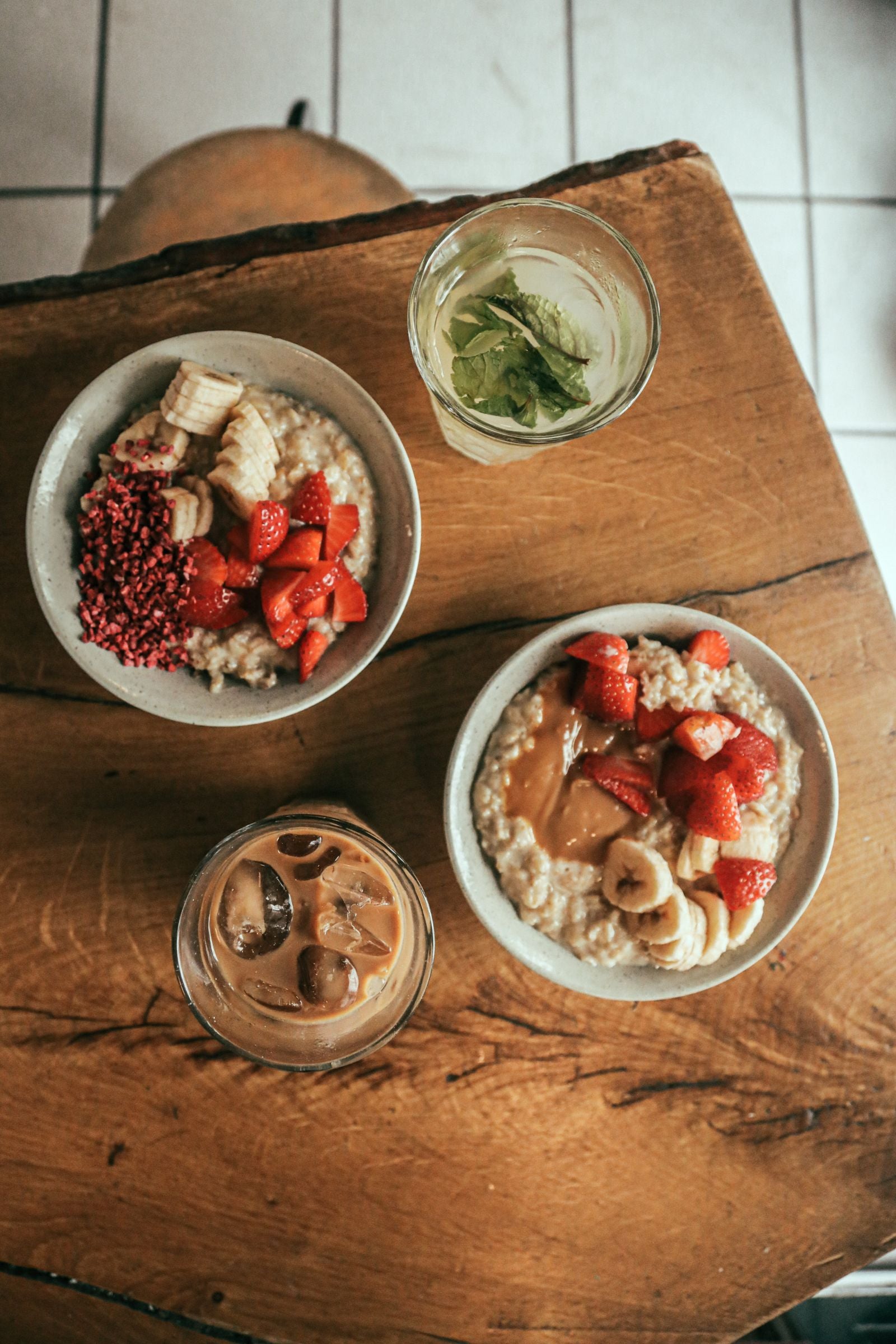 Zwei Keramikschalen mit Haferbrei, Erdbeeren und Banane auf einem alten Holztisch, daneben Eiskaffee und Wasser mit Minze, von oben im Tageslicht
